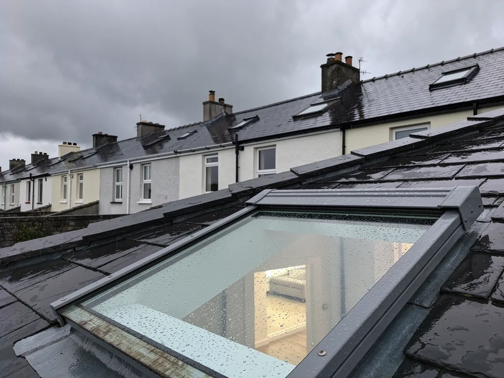 A row of Welsh render terraces with roof windows in wet slate roofs, low grey cloud