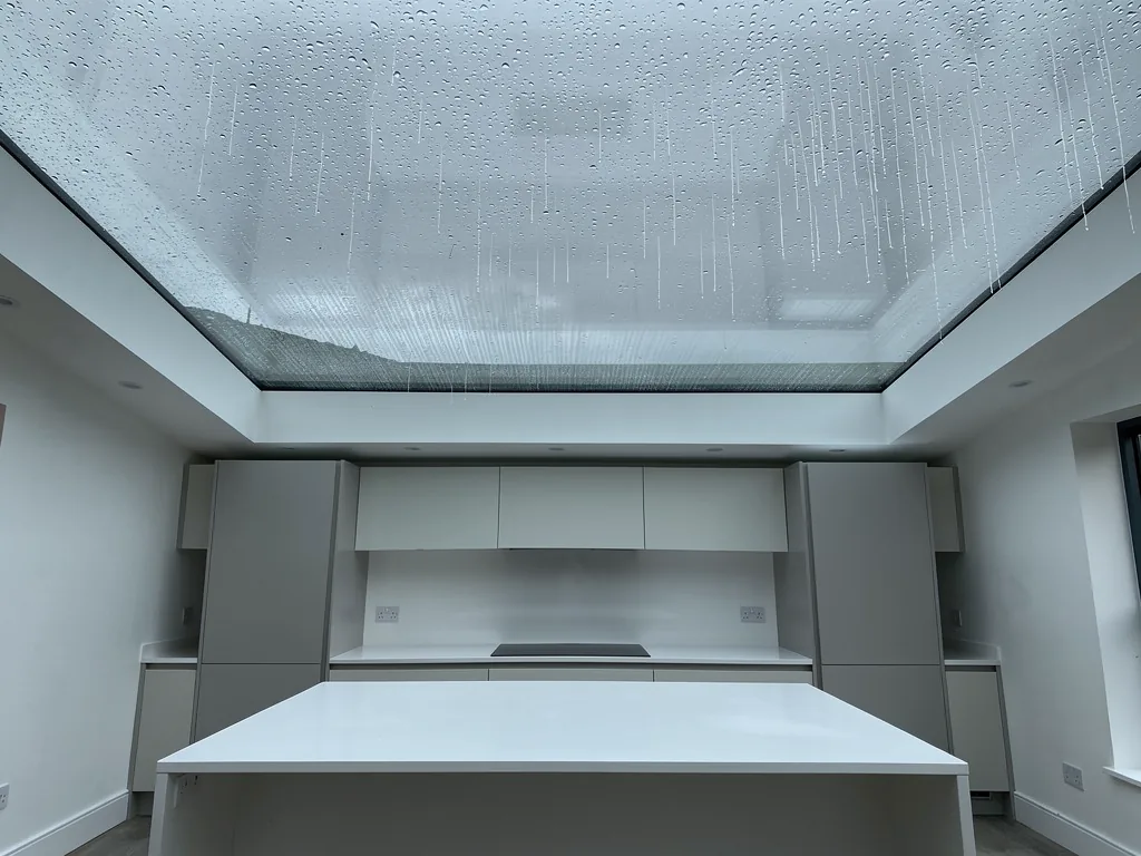 Rain running down a large flat rooflight above a bright white kitchen island, grey sky
