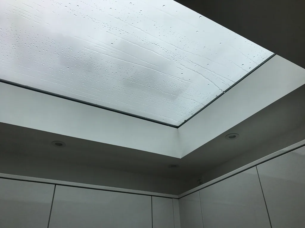 Looking up through a new flat rooflight in a bright kitchen, rain on the glass, grey sky