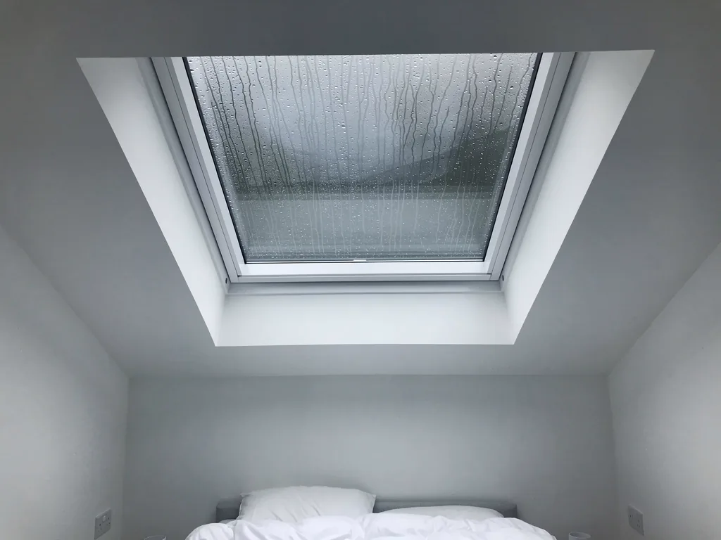 A roof window seen from inside a white loft bedroom, rain running down the pane