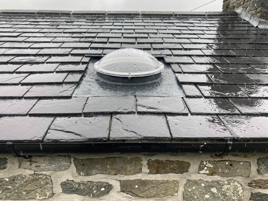 A small round clear sun tunnel dome on a wet slate roof above an old stone wall