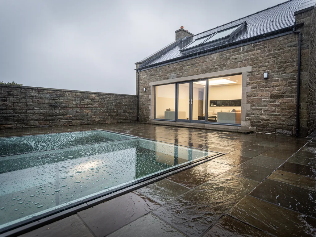 A large walk-on glass panel in a wet patio beside a stone house, rain beading